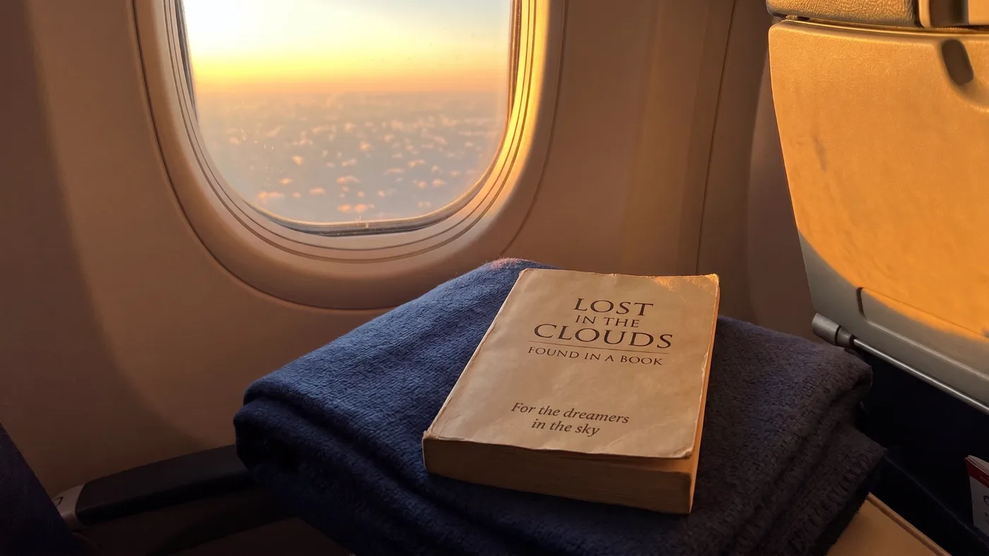 A worn paperback resting on a folded navy blanket beside an airplane window at sunset, with clouds below